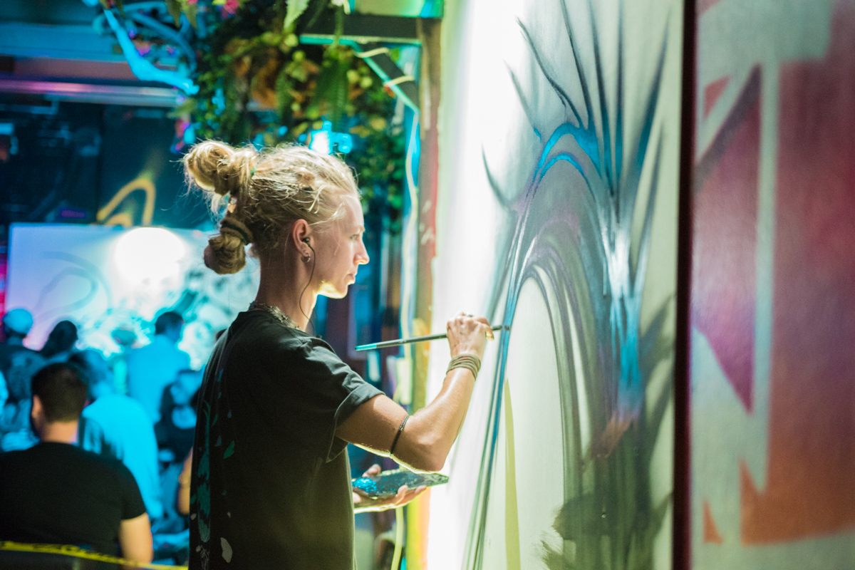 An artist with dreadlocks painting a flowing design on a wall at night