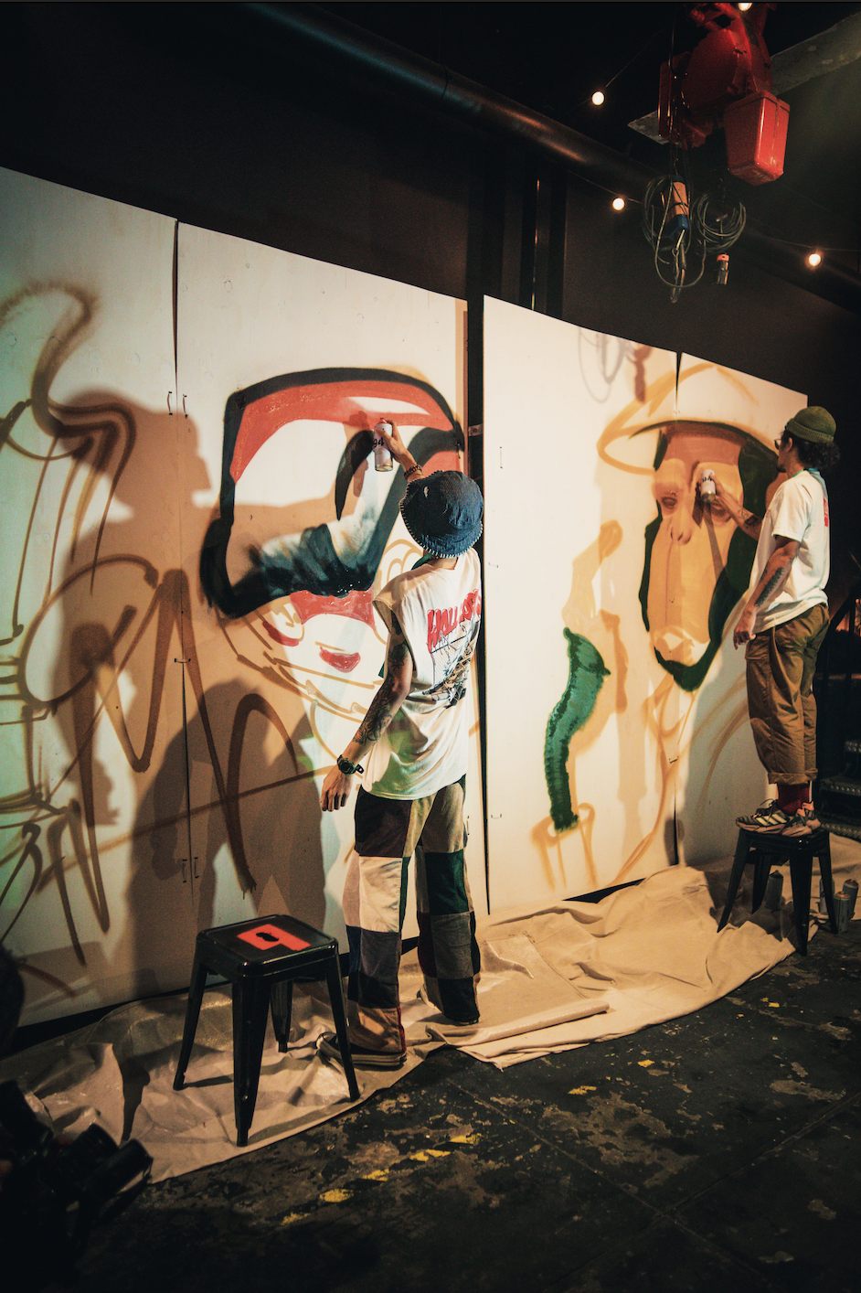 An artist reaching up to paint an expressive red-and-cream face on a tall panel