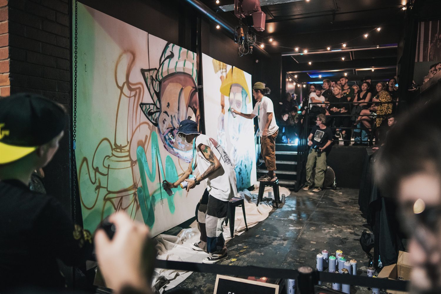 Two artists painting large canvases side by side as a crowd watches at a Scribble Slam battle