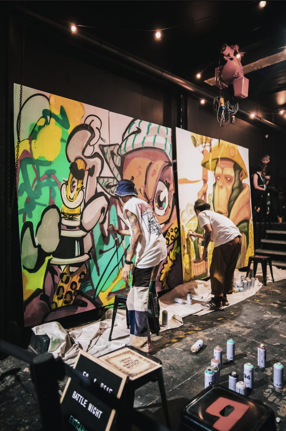 Two artists spray-painting colourful graffiti characters beside a hand-lettered Battle Night sign and rows of spray cans