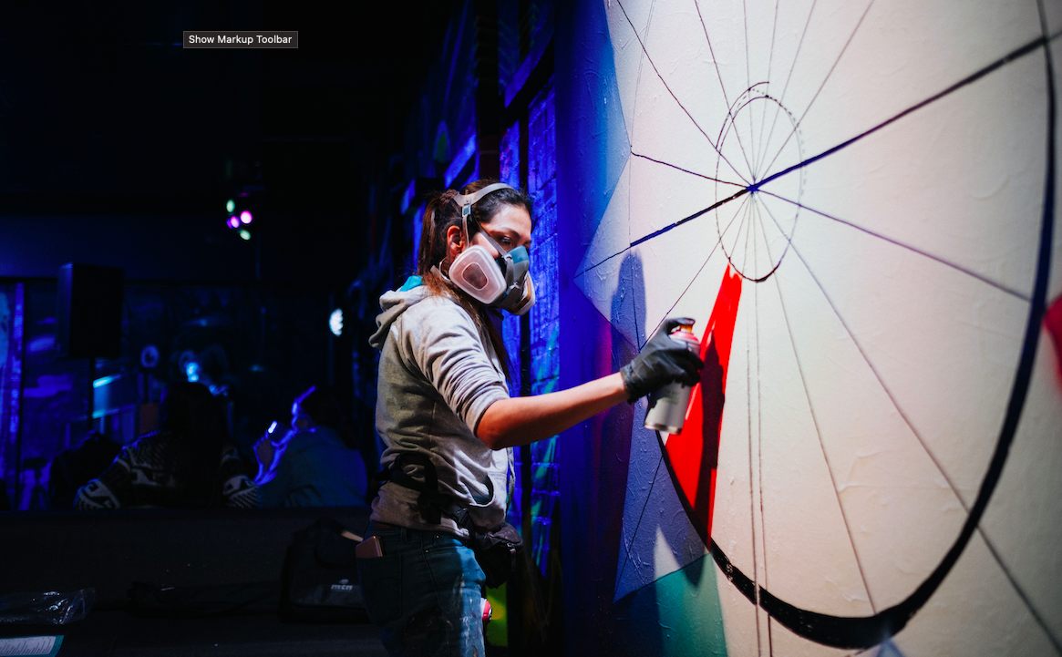 A masked artist in a respirator spray-painting a geometric piece under dramatic blue light
