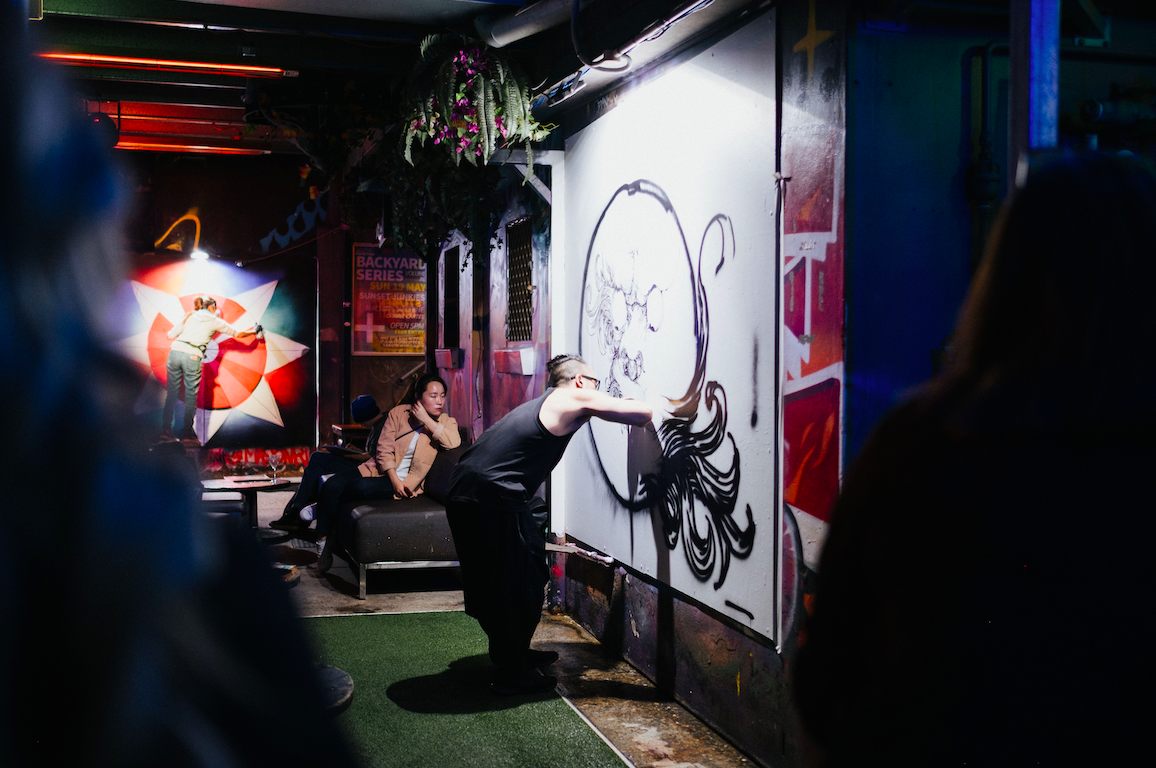 An artist mid-motion painting a black octopus on a white panel in a dim bar